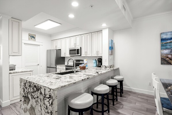 A modern kitchen with white cabinets, a marble island, and a coffee maker. - Gone Coastal - Destin Gulfgate 1103! (Destin)