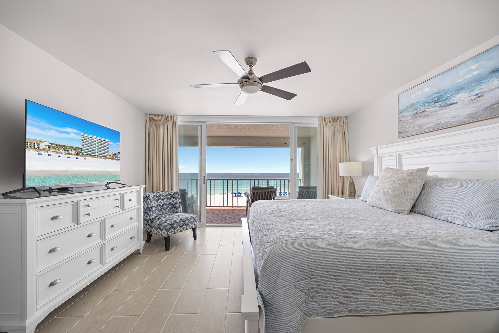 A hotel room with a bed, a TV, a ceiling fan, a balcony with a view of the beach, and a painting on the wall.