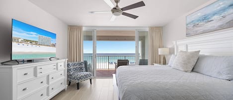 A hotel room with a bed, a TV, a ceiling fan, a balcony with a view of the beach, and a painting on the wall.