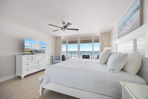 A hotel room with a bed, a ceiling fan, a TV, a balcony with a view of the ocean, and a painting on the wall. - Shore Beats Work - Destin Gulfgate 0201! (Destin)