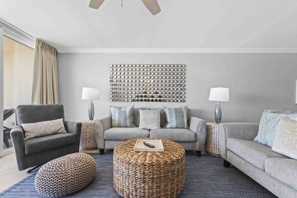 A modern living room with a grey sofa set, a grey armchair, a round wicker ottoman, and a decorative wall art piece.