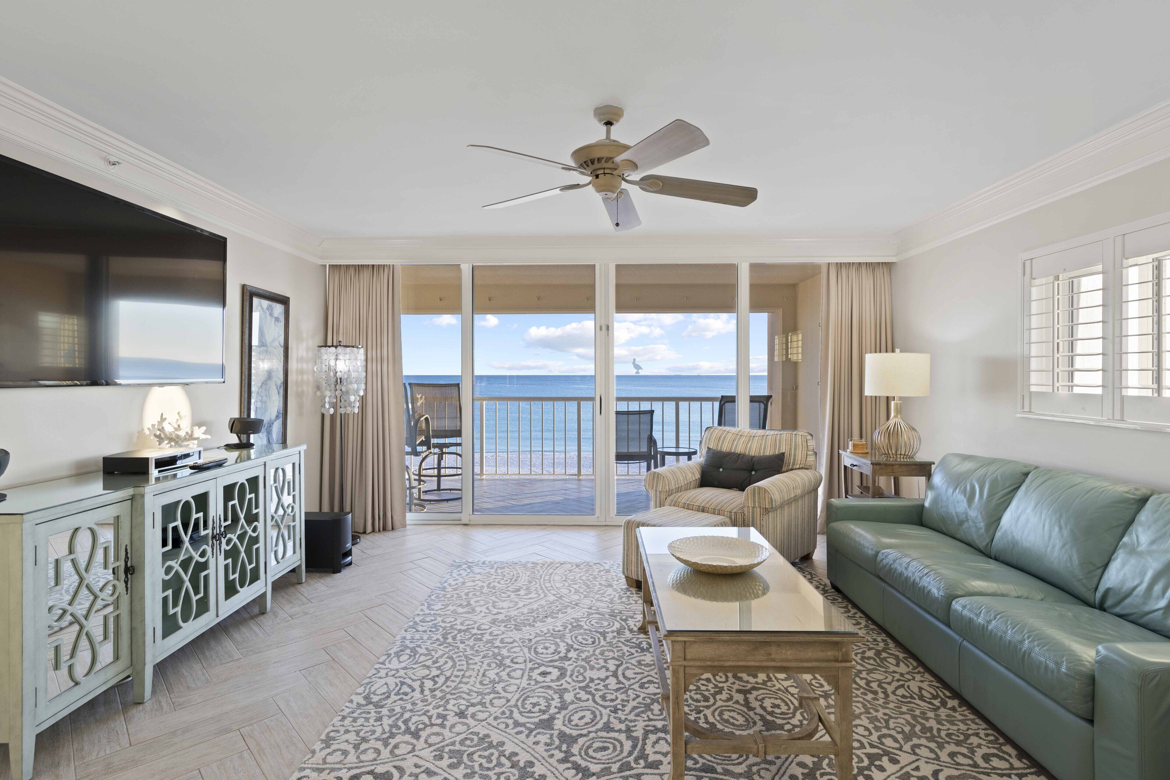 A spacious living room with a green sofa, a coffee table, and a television. There is a balcony with a view of the ocean.