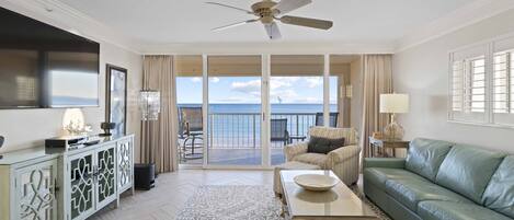 A spacious living room with a green sofa, a coffee table, and a television. There is a balcony with a view of the ocean.