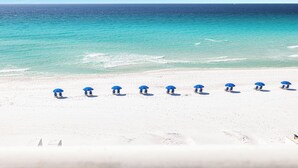 A beach with white sand and a clear blue sea. There are several blue umbrellas and lounge chairs arranged in a row. - Reel Paradise - Destin Gulfgate 0505! (Destin)