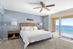 A bedroom with a large bed, a ceiling fan, a nightstand with a lamp, a wall-mounted fish decoration, and a view of the ocean through a sliding glass door. - Absolute Seaclusion - Destin Gulfgate 0506! (Destin)