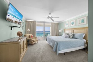 A hotel room with a bed, a chair, a desk, a TV, and a balcony with a view of the ocean.