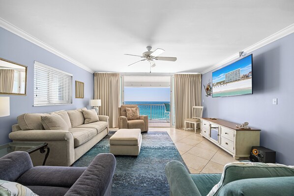 A spacious living room with a balcony, a ceiling fan, a television, and a view of the ocean.