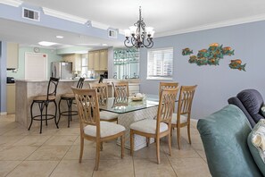 A dining area with a glass table and wooden chairs, a kitchen in the background, and a decorative wall piece.