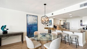A modern kitchen with a central island, a dining area with a glass table and chairs, and a large abstract painting on the wall. - Where every hour is Happy Hour - Destin Gulfgate 0602! (Destin)