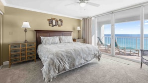 Een slaapkamer met een groot bed, een plafondventilator, een nachtkastje met een lamp en uitzicht op de oceaan door een schuifdeur.