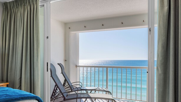 A balcony with a view of the ocean, a bed with a blue blanket, and a chair.