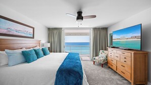 A hotel room with a bed, a TV, a wooden dresser, and a balcony with a view of the ocean.