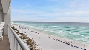 Ein Balkon mit weißen Stühlen mit Blick auf einen Strand, auf dem sich Personen und Sonnenschirme befinden.