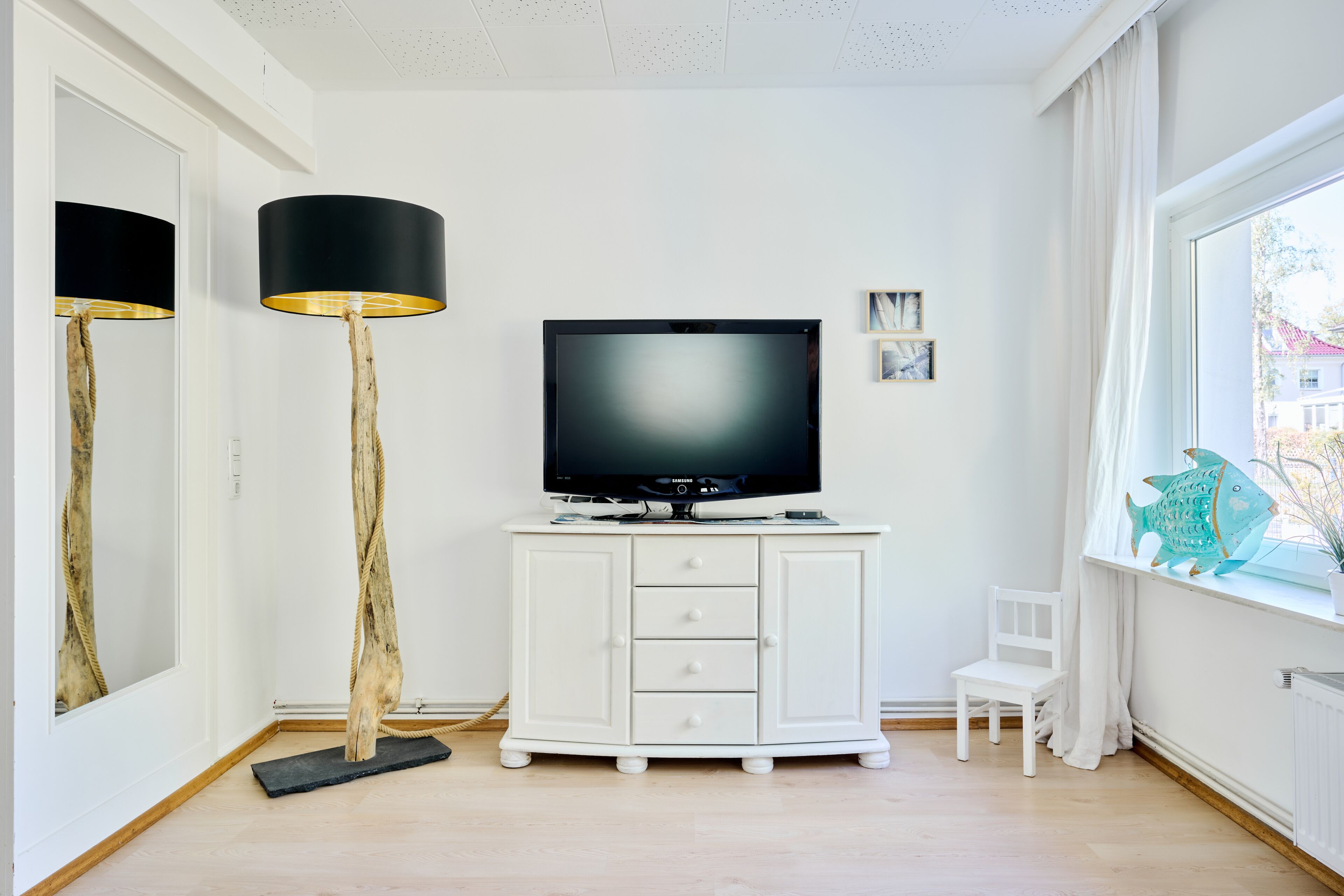 A room with a white cabinet, a flat-screen TV, a tall floor lamp, a small white chair, and a window with a view of buildings.