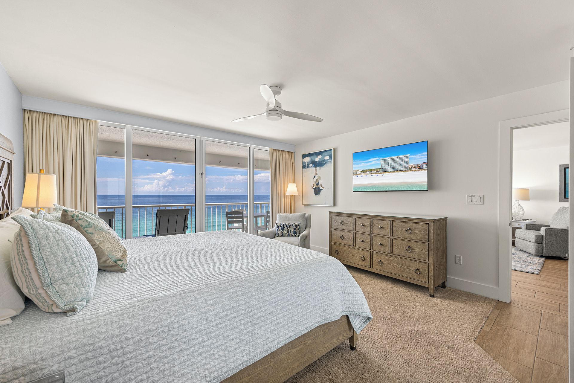 A bedroom with a bed, a dresser, a ceiling fan, and a large window with a view of the ocean.