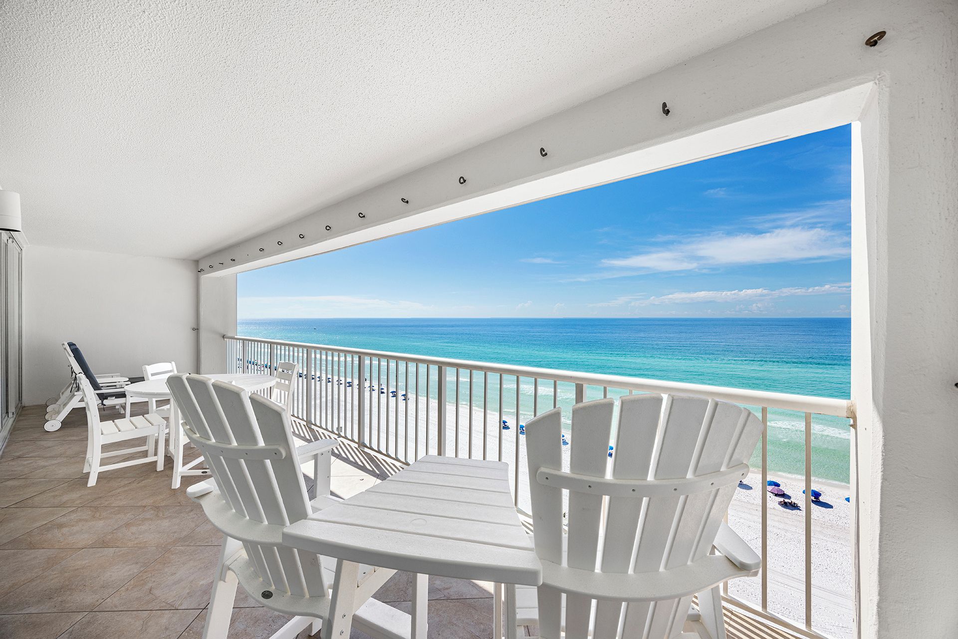 A balcony with white chairs and a table, overlooking a beach with clear blue water.