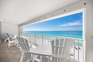 Un balcon avec des chaises blanches et une table, donnant sur une plage aux eaux d’un bleu limpide.