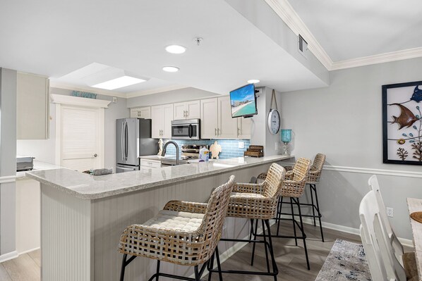 A modern kitchen with a central island, bar stools, a flat-screen TV, and a clock on the wall.