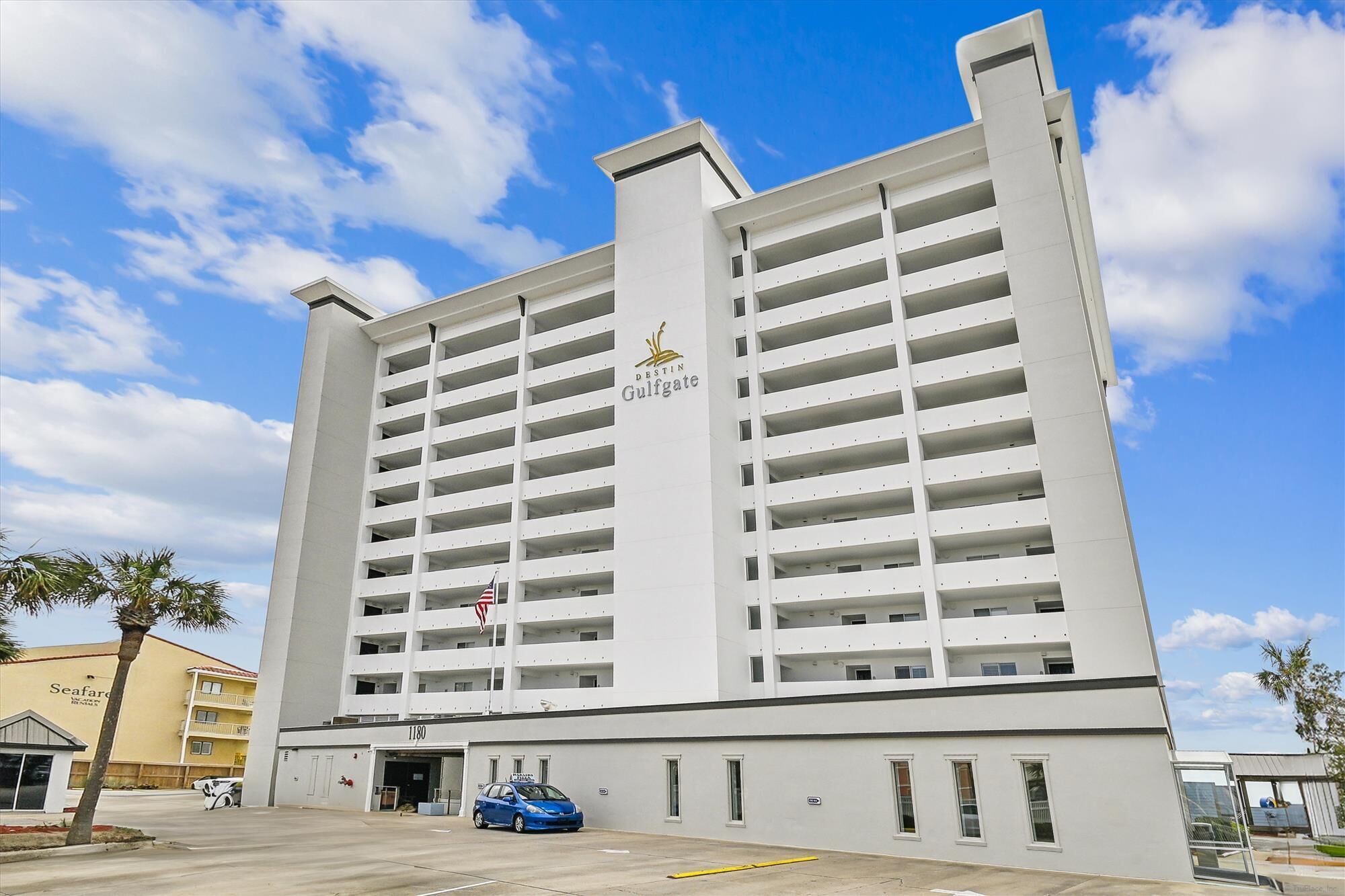 A high-rise building with multiple balconies, a prominent 'Destin Gulfgate' sign, and a parking lot with a blue car.