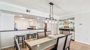 A modern kitchen with a central island, a dining area with a glass-top table, and a pendant light fixture. - All Decked Out - Destin Gulfgate 0904! (Destin)