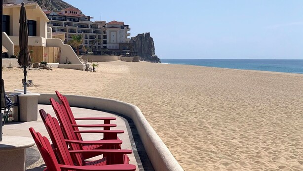 Une plage avec des chaises rouges et un grand parasol, une falaise rocheuse et des bâtiments en arrière-plan.