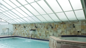 Una piscina interior con techo de cristal, pared de piedra y una pequeña cascada.