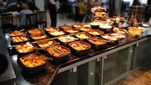 Un bufé con diferentes alimentos en cestas, una partición de vidrio y gente de fondo.