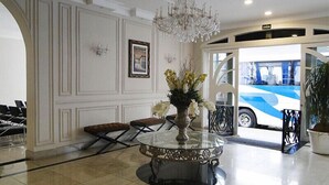 Un vestíbulo con una araña, una mesa redonda con un jarrón de flores, un cuadro en la pared y un autobús fuera de la entrada.