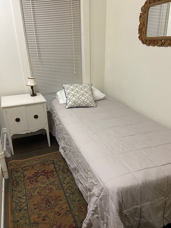 Un dormitorio con una cama individual, una mesita de noche con lámpara, un cojín estampado, una ventana con persianas y un espejo en la pared.