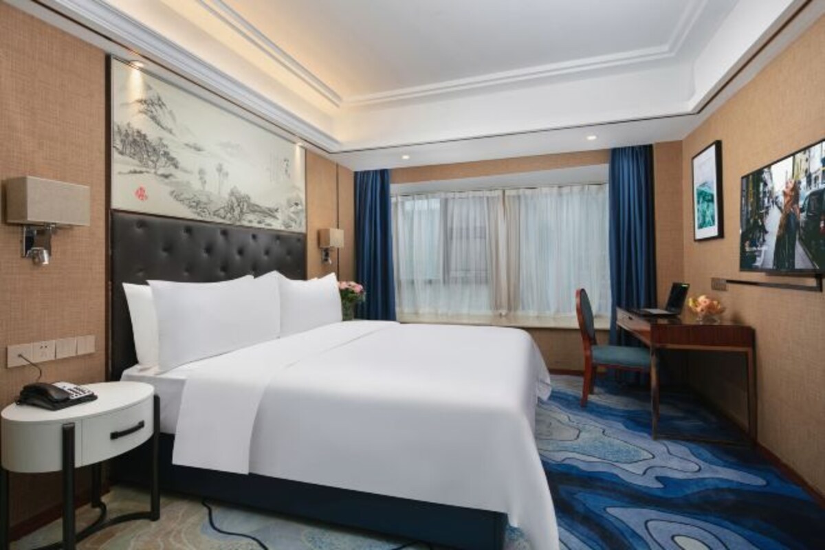 A hotel room with a large bed, a desk, a chair, a small table, a wall-mounted phone, and a painting on the wall.