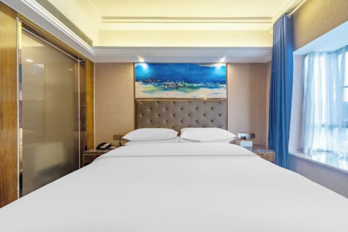A hotel room with a large bed, a headboard with a painting, a nightstand, and a window with curtains.