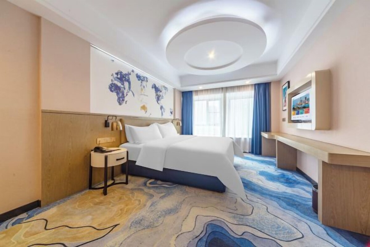A hotel room with a large bed, a desk, a chair, a nightstand, a world map on the wall, and a large window with curtains.