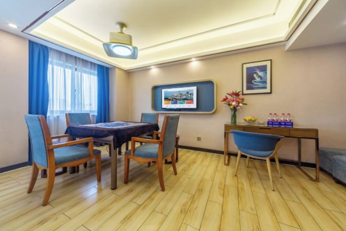A hotel room with a dining area featuring a wooden table and chairs, a television mounted on the wall, and a small side table with a vase of flowers and bottles.
