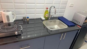 Una cocina con encimera negra, un lavamanos de acero inoxidable, una tetera, una botella de detergente y un microondas.