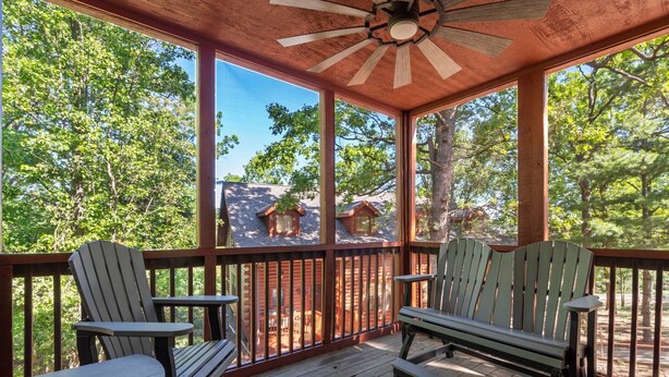 Eine Holzterrasse mit Adirondack-Stühlen und einer Bank, mit Blick auf ein bewaldetes Gebiet, im Hintergrund ein Ferienhaus.