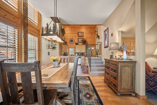 A spacious dining area with a wooden table and chairs, a kitchen in the background, and a staircase leading to another level.