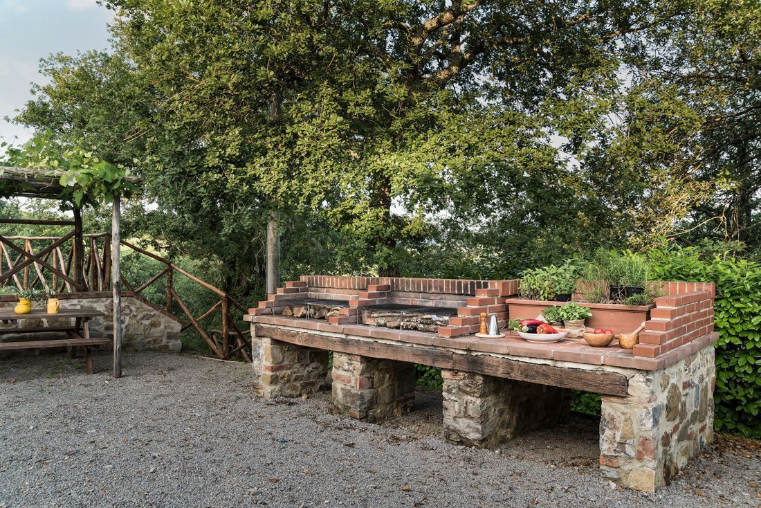 An outdoor brick barbecue area with a stone table, surrounded by greenery and a wooden pergola structure.