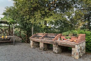 An outdoor brick barbecue area with a stone table, surrounded by greenery and a wooden pergola structure.