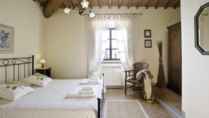Ein Schlafzimmer mit Bett, Nachttisch, Stuhl, einem Fenster mit Vorhängen und einer Holztür.
