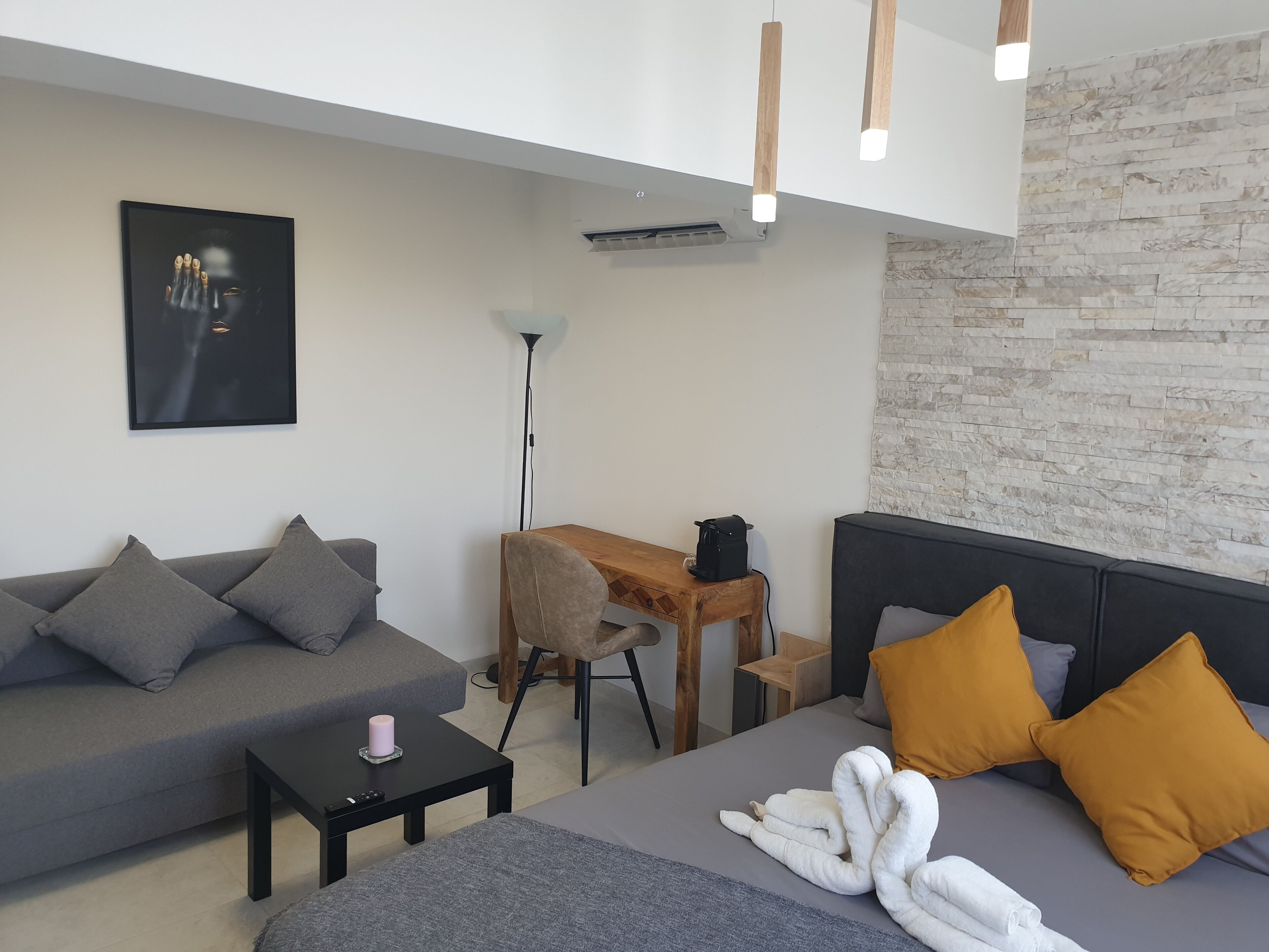 Ein modernes Wohnzimmer mit einem grauen Sofa, einem Holznachttisch, einem schwarzen Couchtisch und einem Bett mit grauer Decke und weißen Handtüchern.