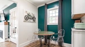 Une pièce aux murs de couleur turquoise, une table ronde en verre, deux chaises en plastique transparent, une œuvre d’art représentant une moto accrochée au mur et une fenêtre avec des rideaux.