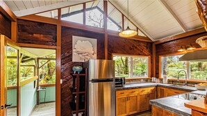A spacious kitchen with wooden cabinets, a stainless steel refrigerator, and large windows offering views of the surrounding forest. - Darling, Private Cottage Near Hawaii Volcanoes Nat'l Park (Volcano)