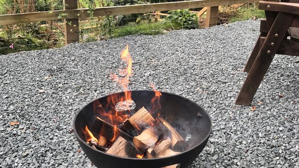 A fire pit with burning wood surrounded by gravel, a wooden fence, and a grassy landscape with flowers in the background. - Shepherds Hut, self catering, Powys (Cascob)