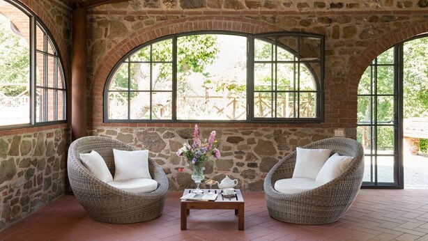 A stone-walled room with two wicker chairs, a small table with a vase of flowers, and large arched windows.