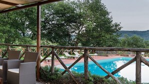 A wooden deck with wicker chairs overlooking a swimming pool and lounge chairs, surrounded by trees.