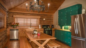 A wooden cabin interior with a dining table, bench, wooden floors, a stainless steel refrigerator, and teal cabinets.