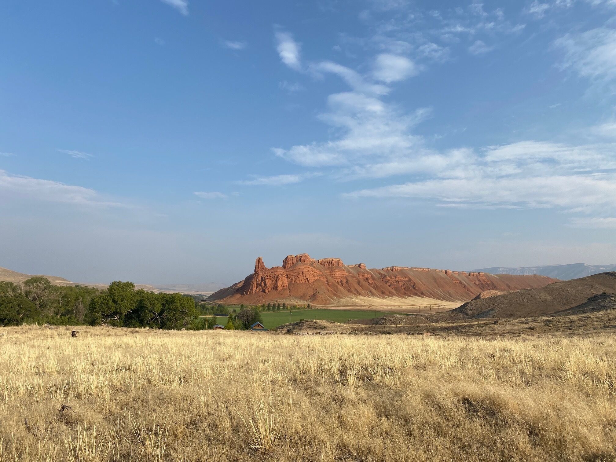 A vast landscape with a prominent red rock formation in the distance, surrounded by dry grass fields and sparse vegetation.