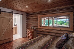 Un dormitorio tipo cabaña de madera con cama, cómoda, ventana con persianas y puerta que da a un baño.