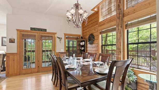 A dining room with a wooden table set for six, a chandelier, and large windows with a view of greenery.
