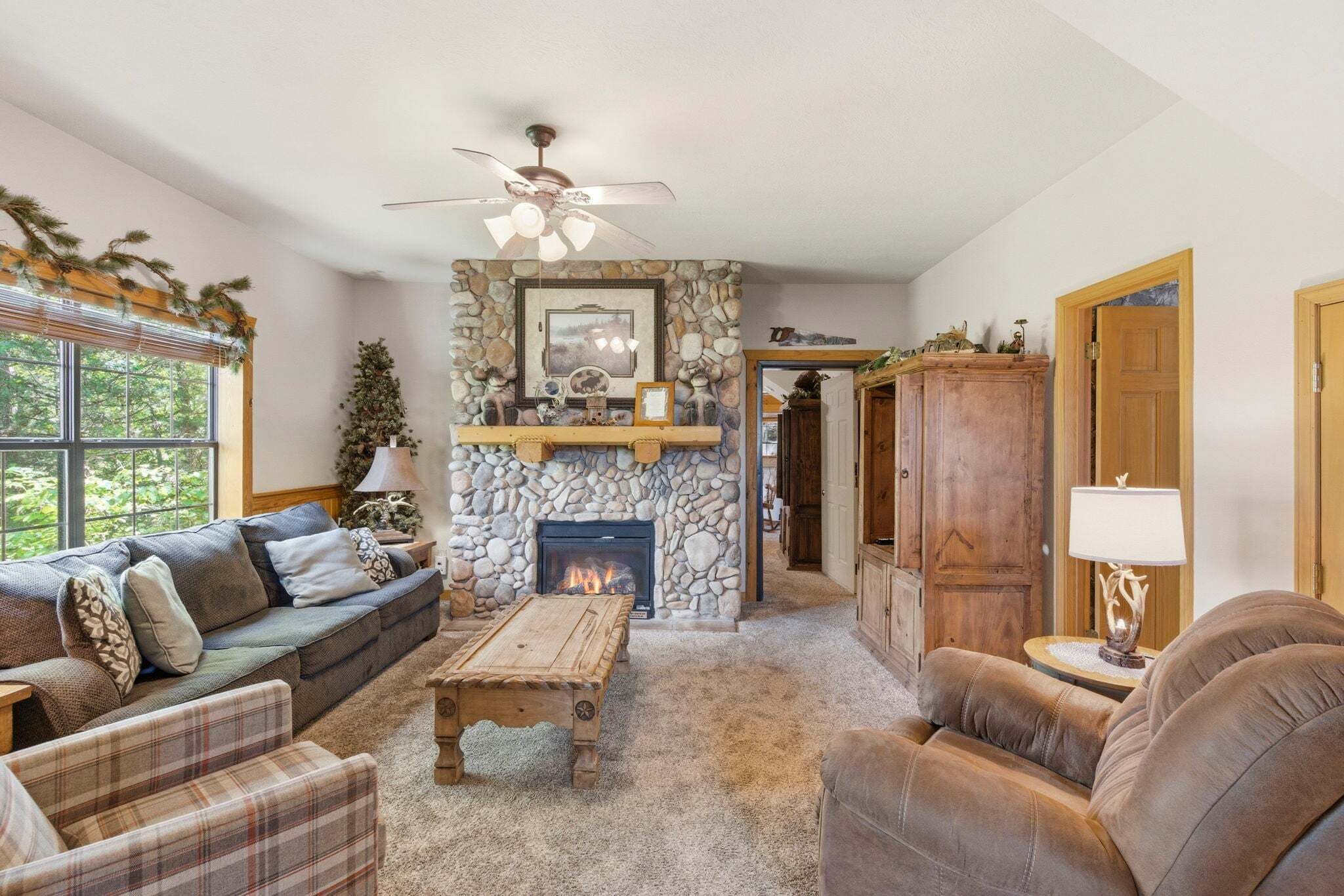 A cozy living room with a stone fireplace, a ceiling fan, and a large window.
